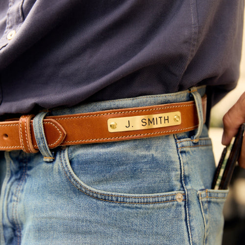 A close-up of a person wearing a brown leather belt with a custom engraved gold metal nameplate that reads 'J. SMITH', paired with a navy blue button-down shirt and light blue jeans.
